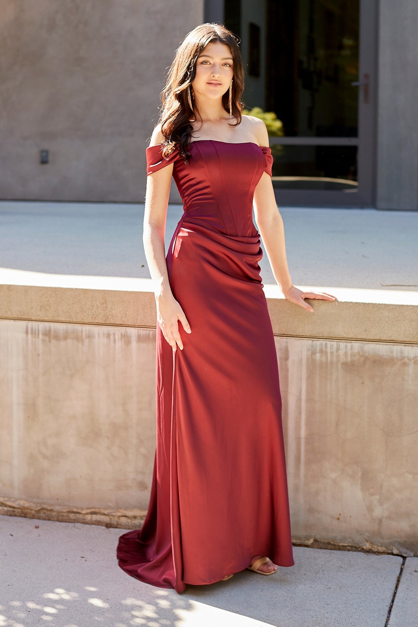 A woman wearing an Amelia Couture 6151 floor-length, off-the-shoulder burgundy gown with a corset-style bodice.